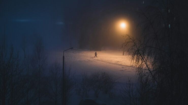A lone pedestrian walking along a snowy road at night under a street lamp